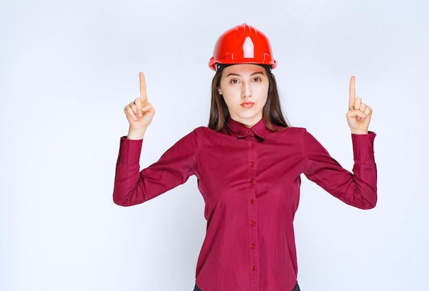 Confident female architect in red hard helmet standing and posing over white wall. 
