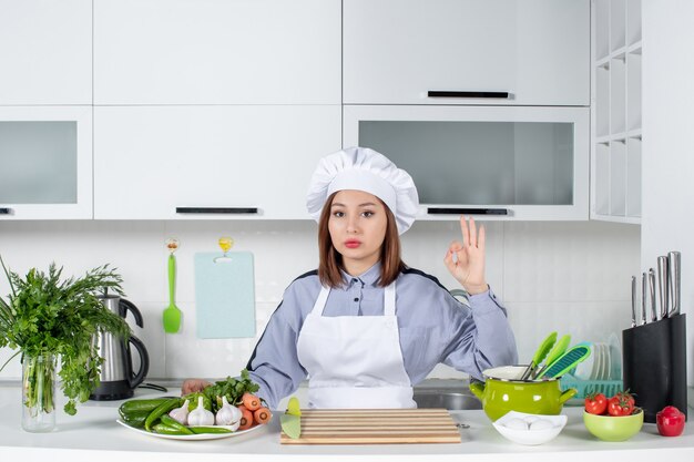Бесплатное фото Уверенная женщина-шеф-повар и свежие овощи с кухонным оборудованием и жестом в очках на белой кухне
