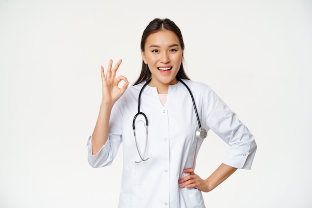 자신감 있는 여성 의사, 의료 유니폼과 청진기를 입은 아시아 의사, 괜찮은 표시를 보여주고 기쁘게 끄덕이며 칭찬하고 좋은 흰색 배경을 추천합니다.