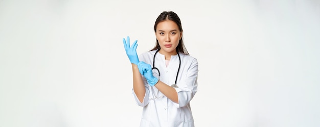 Уверенная в себе женщина-врач-медсестра надела резиновые медицинские перчатки для клинического осмотра пациента, стоящего серьезно в униформе медицинского работника на белом фоне