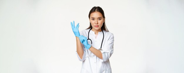 Confident female nurse physician put on rubber medical gloves for patient clinical examination standing serious in healthcare worker uniform white background