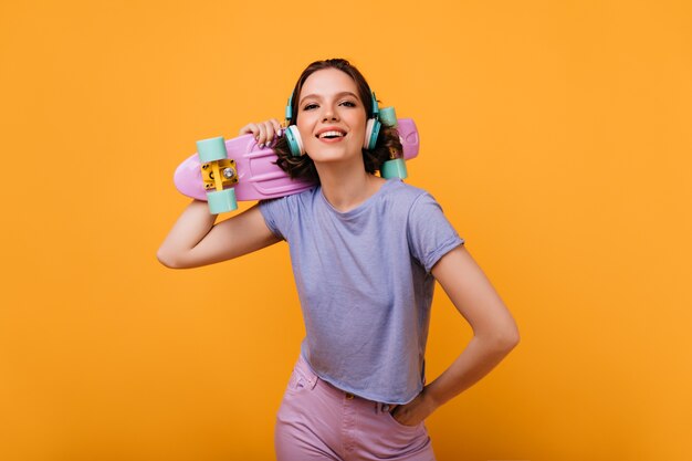 Confident female skateboarder looking with interested smile. pleasant brown-haired woman in headphones isolated.