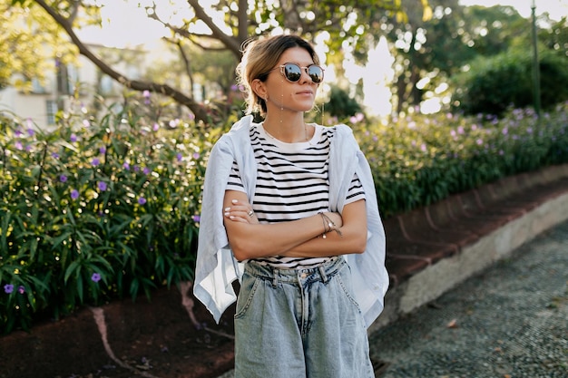 Confident girl in stylish summer outfit wearing sunglasses with charming smile resting outdoor sunny day Portrait of joyful young woman waiting for friends in city garden