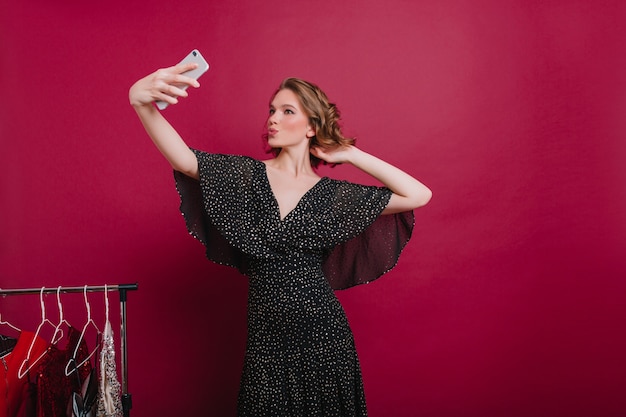 Confident girl with kissing face expression making selfie in her dressing room. stylish woman taking picture of herself near clothes hangers.