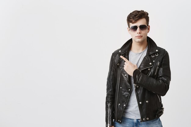 Free photo confident good-looking guy in black leather jacket with sunglasses on, indicates with fore finger at copy space for advertisment or promotional text. handsome stylish male points into distance