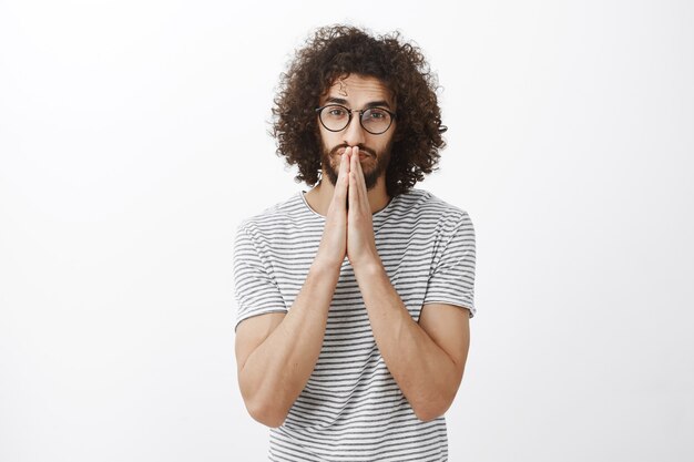 Confident good-looking hispanic guy with curly hair in trendy black glasses, holding hands in pray over mouth and nose