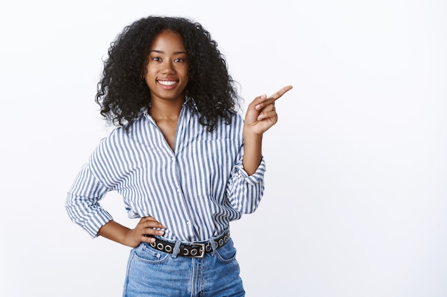 Confident good-looking outgoing nice dark-skinned curly-haired female friend suggesting visit cozy cafe pointing sideways inviting come, smiling broadly friendly vibe, holding hand waist casually