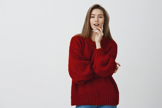 自信を持って見栄えの良い生意気なセクシーなヨーロッパの女性は赤いルーズセーターを噛んで指を大胆で軽薄に見ています。