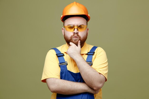 Confident grabbed chin young builder man in uniform isolated on green background