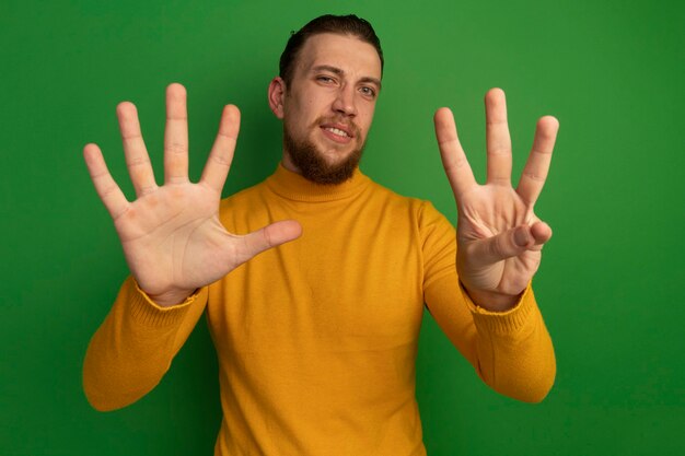 Confident handsome blonde man gestures eight with fingers isolated on green wall