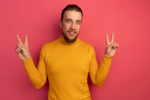 Confident handsome blonde man gestures victory hand sign with two hands on pink