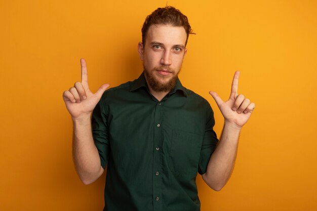 Confident handsome blonde man points up with two hands isolated on orange wall