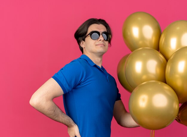 Confident handsome caucasian man in sun glasses stands sideways putting hand on waist and holding helium balloons 