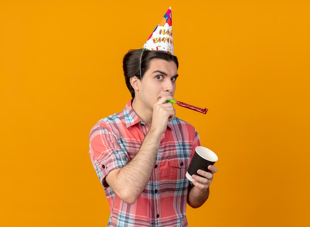 Confident handsome caucasian man wearing birthday cap holding paper cup and blowing party whistle 
