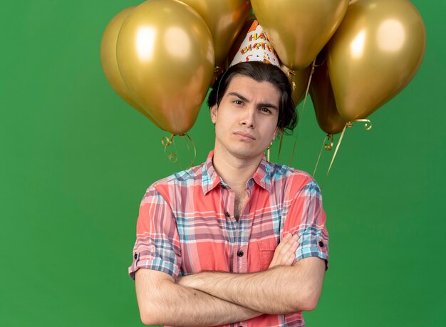 Confident handsome caucasian man wearing birthday cap stands with crossed arms in front of helium balloons looking at camera 