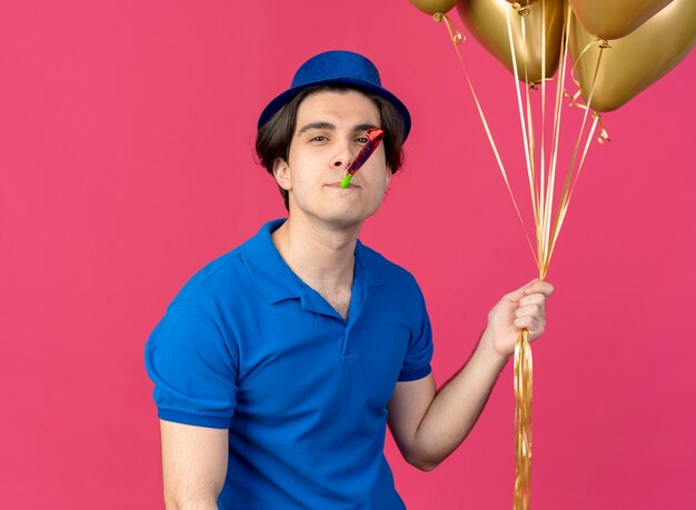 Confident handsome caucasian man wearing blue party hat holds helium balloons blowing party whistle 