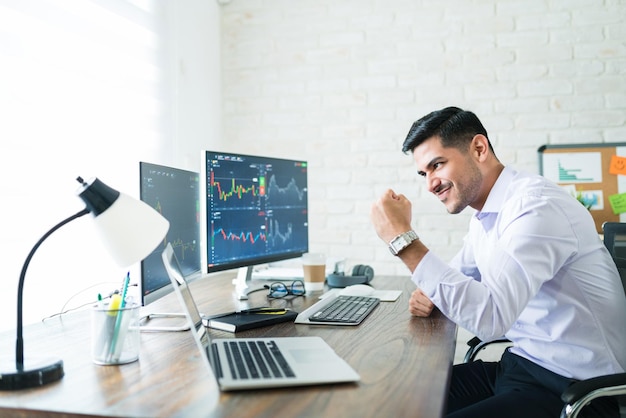 Free photo confident handsome hispanic financial broker showing fist to computer screen while trading from home