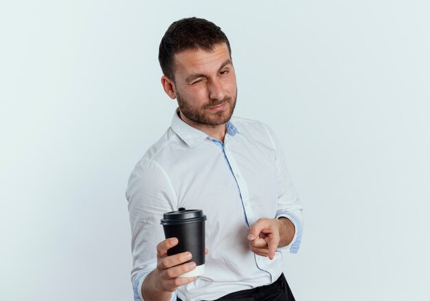 Confident handsome man blinks eye holding coffee cup and pointing isolated on white wall