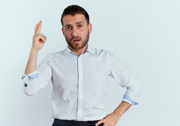 Confident handsome man crosses fingers and puts hand on waist isolated on white wall