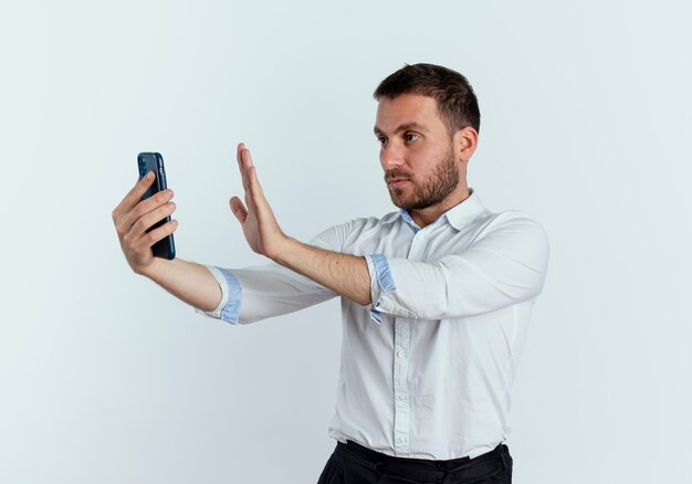 Confident handsome man gestures stop hand sign holding and looking at phone isolated on white wall