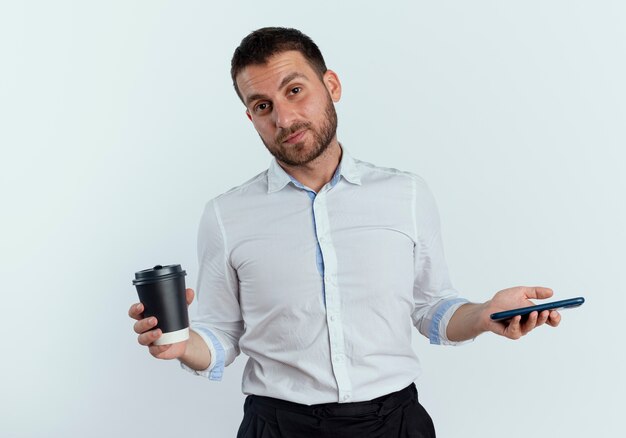 Confident handsome man holds coffee cup and phone isolated on white wall