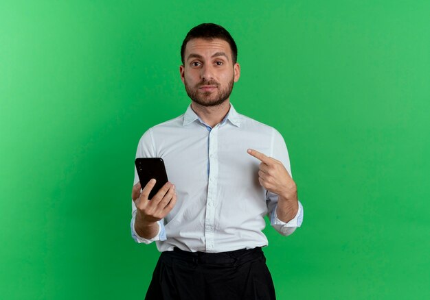 Confident handsome man holds phone and points himself isolated on green wall