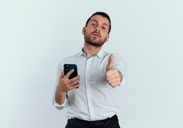 Confident handsome man holds phone and thumbs up isolated on white wall