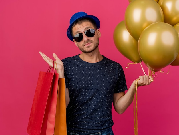 파란색 파티 모자를 쓰고 태양 안경에 자신감이 잘 생긴 남자는 헬륨 풍선과 분홍색 벽에 고립 된 종이 쇼핑백을 보유하고 있습니다.