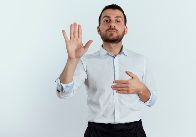 Confident handsome man puts hand on chest and raises hand isolated on white wall