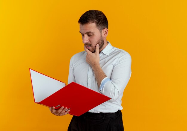 Confident handsome man puts hand on chin looking at file folder isolated on orange wall