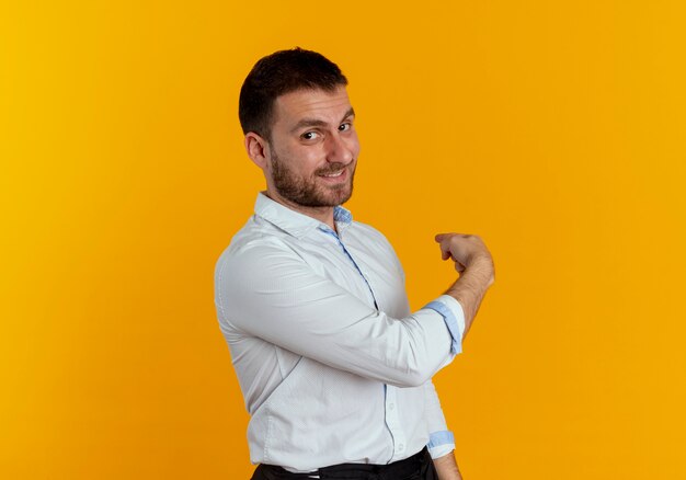 Confident handsome man stands sideways pointing back isolated on orange wall