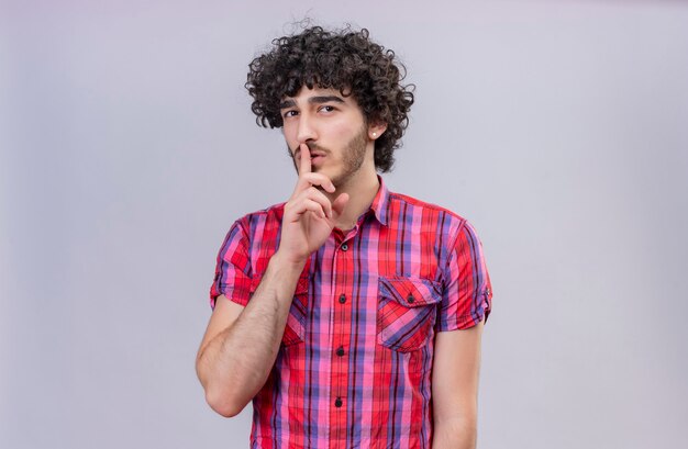 A confident handsome man with curly hair in checked shirt keeping index finger on mouth 