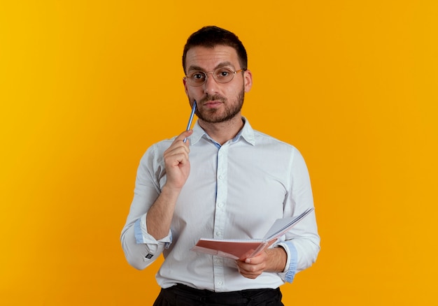 Confident handsome man with optical glasses puts pen on face and holds notebook isolated on orange wall