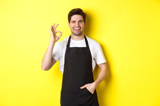 Уверенный и красивый официант показывает знак ОК в черном фартуке и стоит на желтом фоне.
