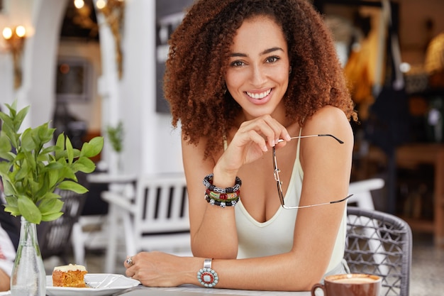 Confident happy dark skinned mixed race young female with curly hair and happy look, dressed casually, recreats in cozy cafe, tastes delicious cake