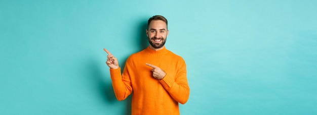 Confident happy guy in orange sweater showing logo banner pointing fingers right at copy space turqu