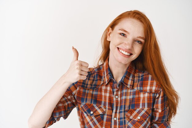 Уверенная и счастливая женщина с длинными рыжими волосами, показывающая большой палец с удовлетворенной улыбкой, одобряет и, как план, согласна с вами или говорит "да" на белом фоне