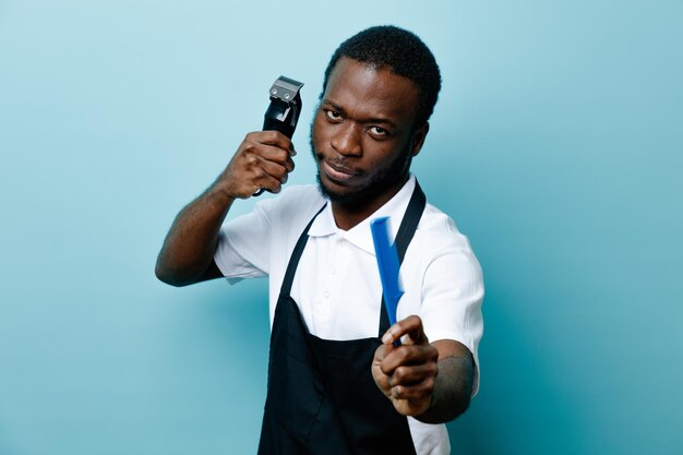 Уверенно держащий расческу с машинкой для стрижки волос молодой африканский парикмахер в униформе, изолированный на синем фоне