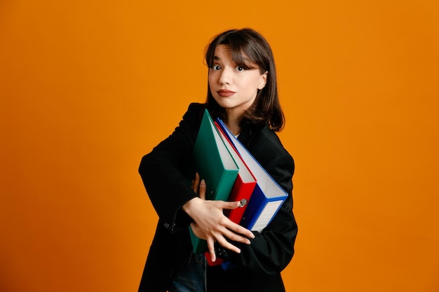 Confident holding folders young beautiful female wearing black jacket isolated on orange background