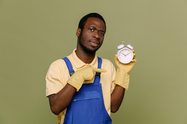 Confident holding and points at alarm clock young africanamerican cleaner male in uniform with gloves isolated on green background