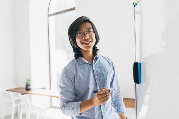 Confident japanese student in trendy glasses holding marker, standing near white board