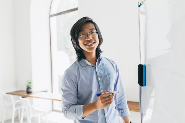 Confident japanese student in trendy glasses holding marker, standing near white board