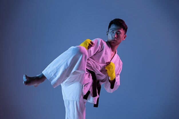 Confident korean man in kimono practicing hand-to-hand combat, martial arts. Young male fighter with black belt training on gradient wall in neon light. Concept of healthy lifestyle, sport.