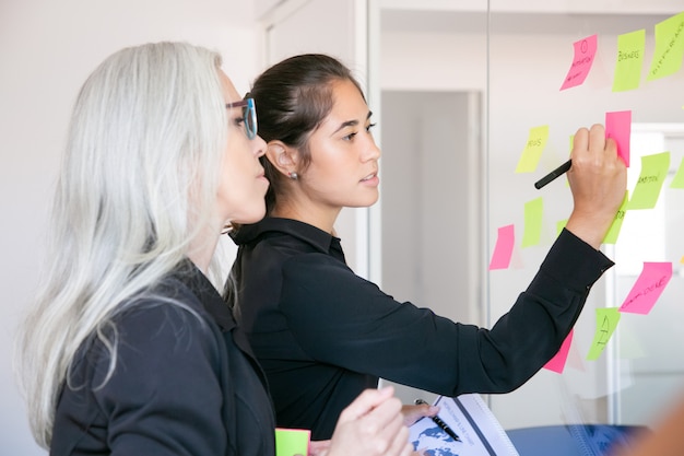 Confident Latin businesswoman writing on stickers and sharing ideas for project. Focused grey-haired female manager reading notes on glass wall