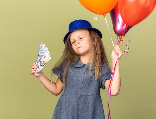 헬륨 풍선과 복사 공간 올리브 녹색 벽에 고립 된 돈을 들고 블루 파티 모자와 자신감이 작은 금발 소녀