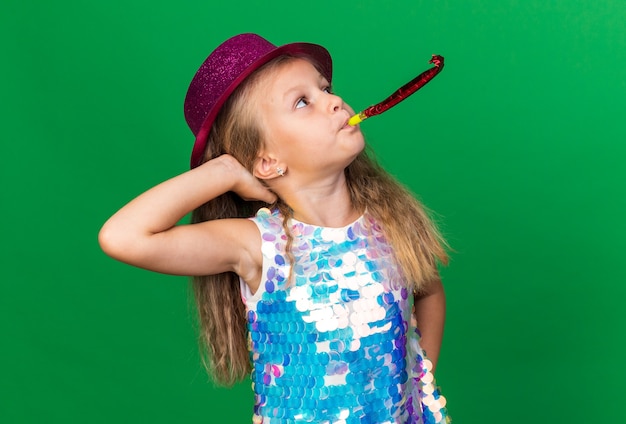 confident little blonde girl with purple party hat blowing party whistle and looking up putting hand on head isolated on green wall with copy space