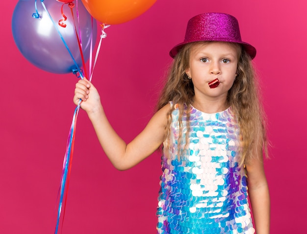 confident little blonde girl with purple party hat holding helium balloons and blowing party whistle isolated on pink wall with copy space