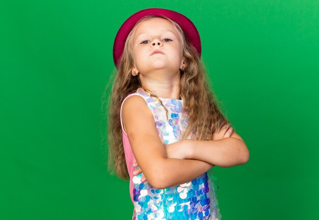 confident little blonde girl with purple party hat standing sideways with crossed arms isolated on green wall with copy space