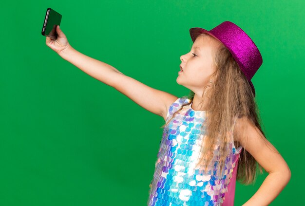уверенная в себе маленькая блондинка в фиолетовой шляпе, делающая селфи на телефоне, изолированном на зеленой стене с копией пространства