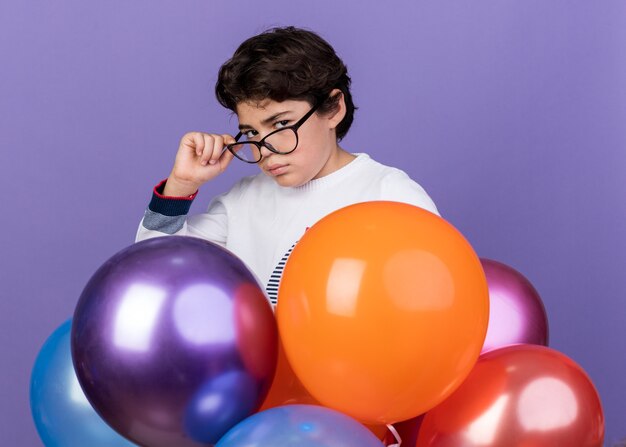 Confident little boy wearing glasses standing behind balloons 